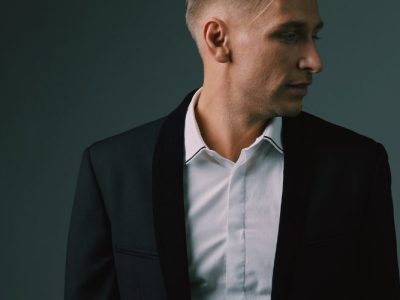 Homem usando terno slim preto com camisa branca, olhando para o lado, em fundo escuro.