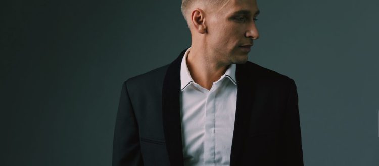 Homem usando terno slim preto com camisa branca, olhando para o lado, em fundo escuro.