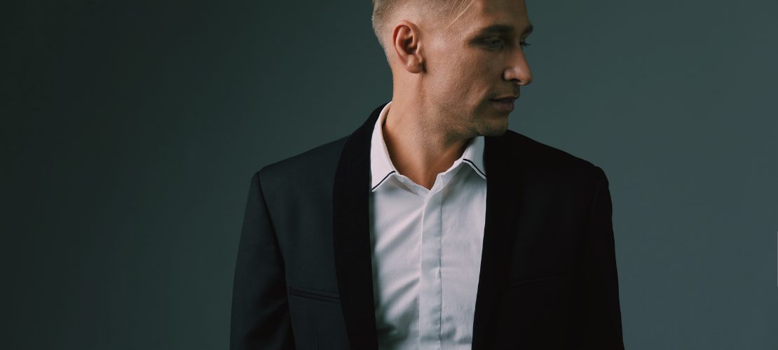 Homem usando terno slim preto com camisa branca, olhando para o lado, em fundo escuro.