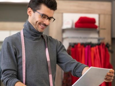 Alfaiate sorrindo em seu ateliê enquanto analisa moldes de roupa sobre uma mesa.