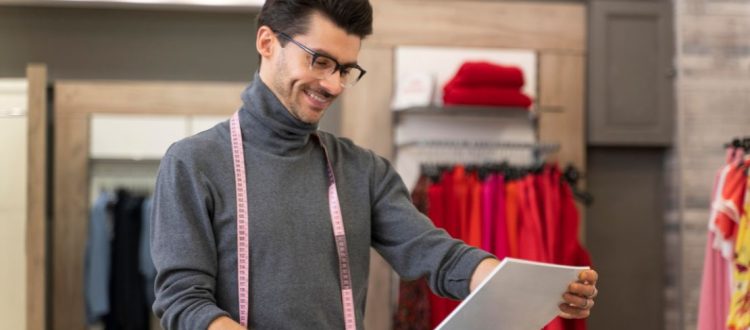 Alfaiate sorrindo em seu ateliê enquanto analisa moldes de roupa sobre uma mesa.