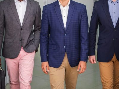 Três homens vestindo blazers sociais em ambiente corporativo, caminhando por um corredor de escritório, com calças em tons neutros e pastas de trabalho.