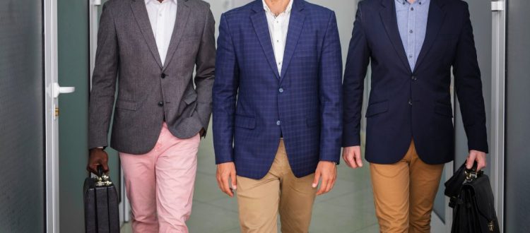 Três homens vestindo blazers sociais em ambiente corporativo, caminhando por um corredor de escritório, com calças em tons neutros e pastas de trabalho.