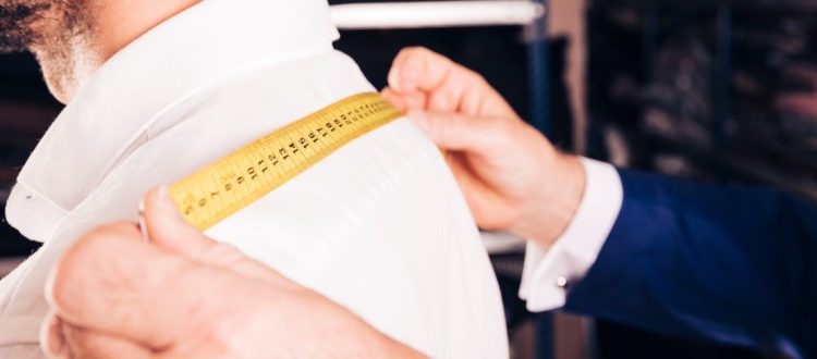 Alfaiate realizando medição dos ombros de um cliente para confecção de roupa sob medida.